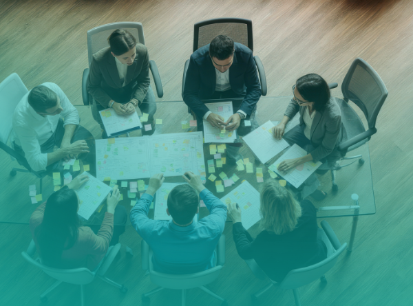 A group of business professionals sit around a glass conference table reviewing documents and sticky notes, collaborating on strategy and planning in a modern office setting.